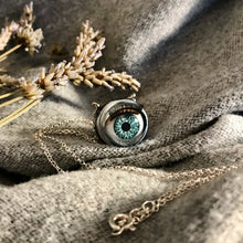 Load image into Gallery viewer, aqua coloured doll eye with metal casing hung on sterling silver chain, item displayed on soft grey fabric with  dried lavender