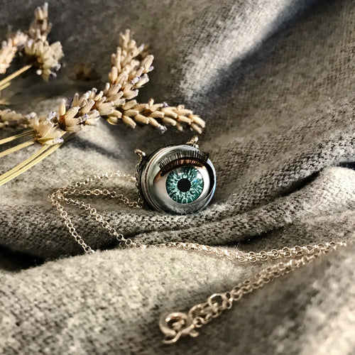 aqua coloured doll eye with metal casing hung on sterling silver chain, item displayed on soft grey fabric with  dried lavender