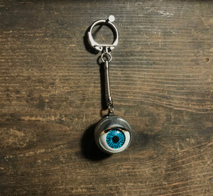 large blue doll eye with metal casing on metal keychain, shown on dark wood background