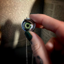 Load image into Gallery viewer, green doll eye with metal casing and long eyelashes held by hand, vintage books are shown in the background