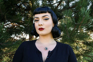 Pin up woman with dark eyemakeup wearing the northern lights eyeball necklace with trees behind 