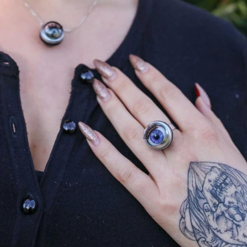 large purple doll eye ring shown on models hand. hand is tattooed and has long glittery nails with hand resting on chest