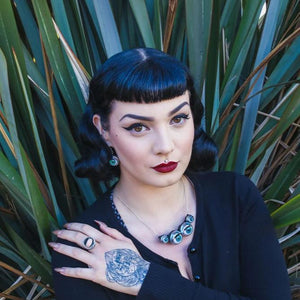 Large necklace made from doll eyes being worn by pin up/gothic model infront of large plant