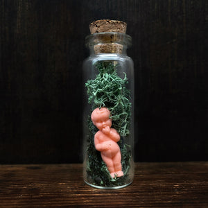 minature glass vial with cork lid, enclosing a very small plastic doll figurine with moss. Doll has a vintage style appearance and is all pink/peach in colour