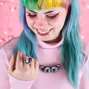 model with rainbow hear and bright pink clothing looks at purple ring, also wearing spider necklace