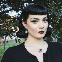 Load image into Gallery viewer, pin up model with black hair and dark eye makeup wears green eyeball necklace and matching earrings in nature with a ewe tree