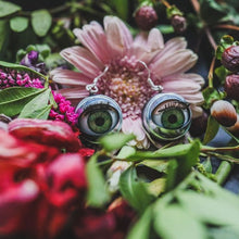 Load image into Gallery viewer, green doll eye earrings with metal casing shown with flowers on dark background