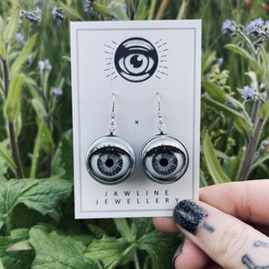 grey doll eye earrings on backing card held infront of forget-me-not flowers