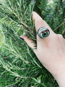 green doll eye ring shown on models hand holding a bunch of rosemary, the iris is a similar colour to the rosemary