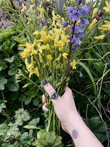 doll eye ring shown worn on thumb, a very green photo with hand holding a bunch of yellow and purple flowers